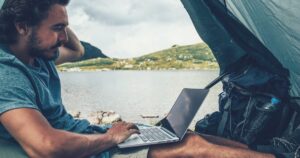 man using a laptop in a tent near a lake