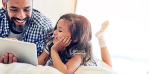 small child laying on bed with father laughing while looking at tablet