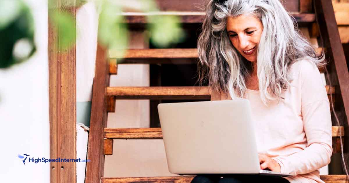 woman with gray hair using laptop on staircase outdoors with tree branch in the front of screen