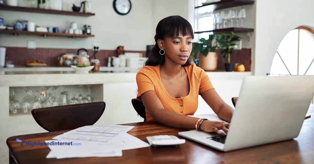 woman wearing orange shirt using laptop at kitchen table with paperwork and calculator next to laptop