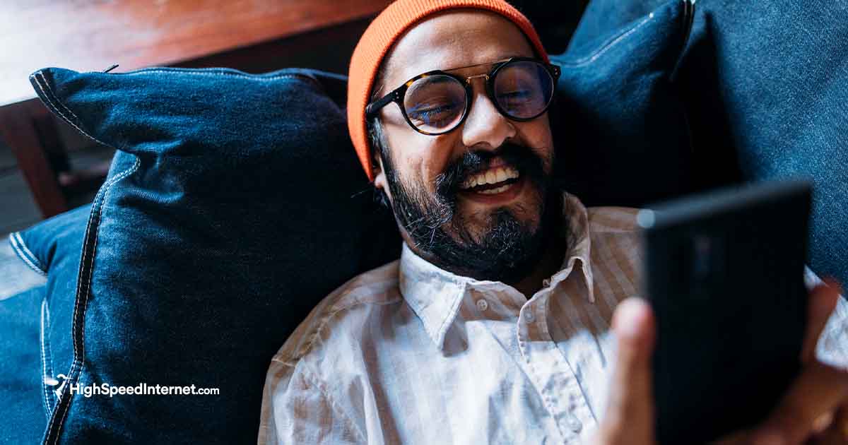 Happy male smiling while using cell phone laying down on a couch. Wearing a yellow knit hat and glasses.