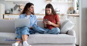 Mother and daughter sitting on couch using cell phone and laptop.