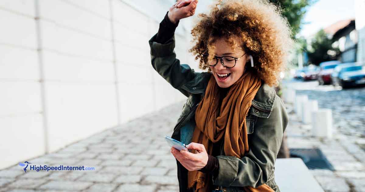 Excited female with an arm in the air and using a cell phone while walking down a cobblestone street.